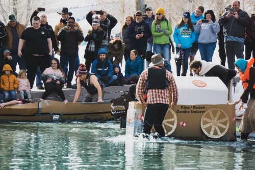 A crowd watches as participants race cardboard boats in a water event, with one boat resembling a dinosaur.