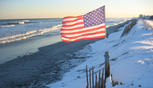 A U.S. flag stands on a snowy beach, with waves gently lapping at the shore under a clear sky.