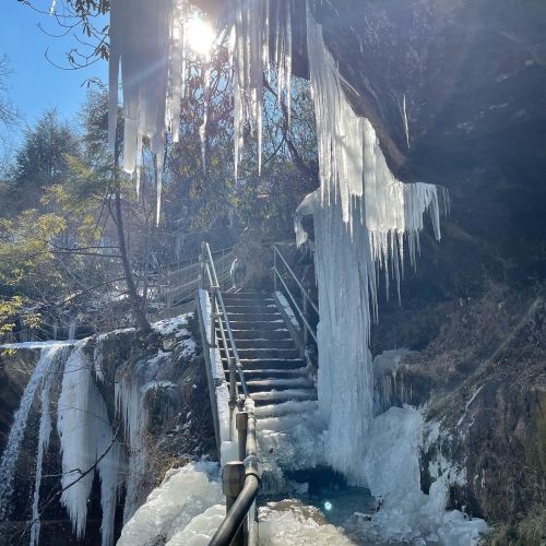 Icicles hang from a rocky overhang near a frozen waterfall, with a staircase leading down to the icy water below.