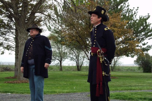 Two men in historical military uniforms stand outdoors, surrounded by trees and grass, with a cloudy sky above.