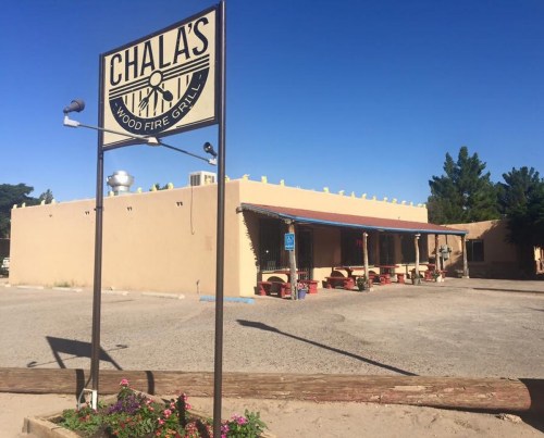 A rustic restaurant building with a sign reading "Chala's Wood Fire Grill," surrounded by gravel and desert landscaping.