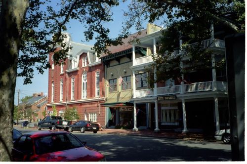 Historic buildings line a street, featuring a red brick structure and a white balcony-adorned facade under a clear blue sky.