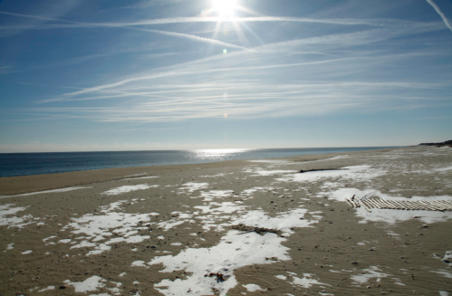 A serene beach scene with snow patches, under a bright sun and clear blue sky, reflecting on the calm ocean.