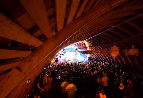 A lively crowd enjoys a concert inside a rustic, wooden barn with beams and colorful stage lights.