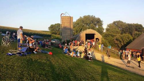 A crowd gathers outside a barn and silo on a grassy hill, enjoying a sunny day at a rural event.