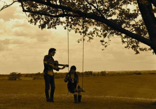 A sepia-toned image of a person playing guitar while another sits on a swing under a tree in a grassy field.