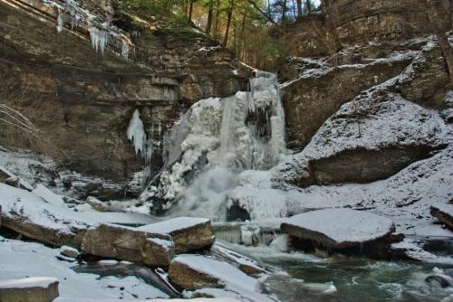 A frozen waterfall surrounded by snow-covered rocks and trees in a winter landscape.