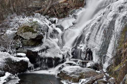 A serene waterfall cascades over icy rocks, surrounded by a winter landscape with frosted vegetation.