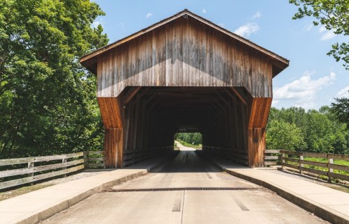 A wooden covered bridge with a clear sky and trees in the background, leading to a paved road.