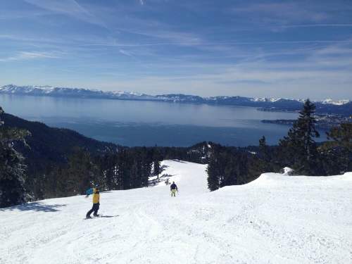 Two skiers descend a snowy slope with a view of a lake and mountains in the background under a clear blue sky.