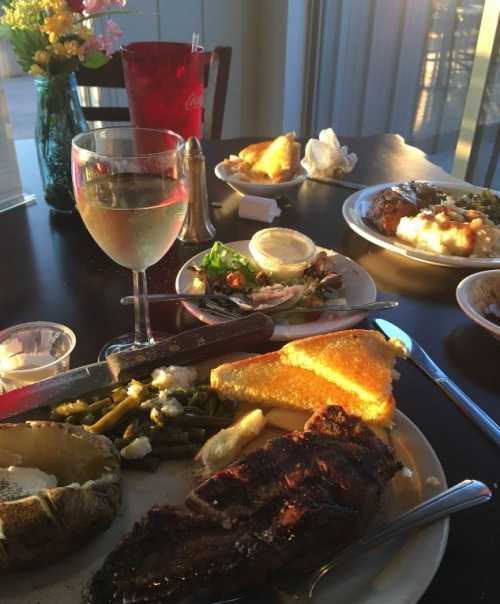 A table set with a glass of white wine, a plate of grilled steak, sides of green beans, cornbread, and a salad.