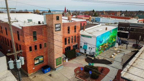 Aerial view of a brick building labeled "Art Loft" next to a colorful mural on a nearby structure in a small town.