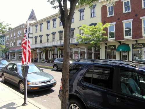 A street view featuring parked cars, storefronts, and an American flag on a pole. Trees line the sidewalk.