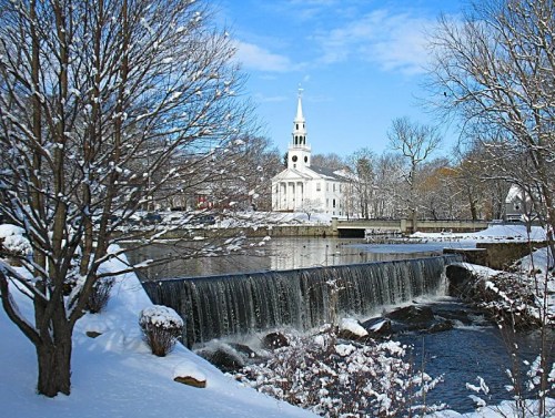 A snowy landscape featuring a church, a waterfall, and trees, under a clear blue sky.