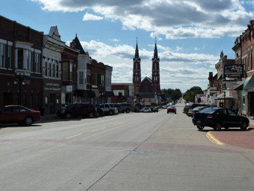 A quiet street lined with shops, leading to a church with twin spires under a partly cloudy sky.