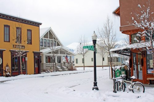 Snow-covered street scene with colorful buildings, a lamppost, and a bicycle parked near a corner sign.