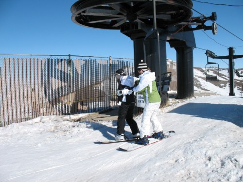 Two skiers at a ski lift, one assisting the other as they prepare to descend a snowy slope. Clear blue sky in the background.