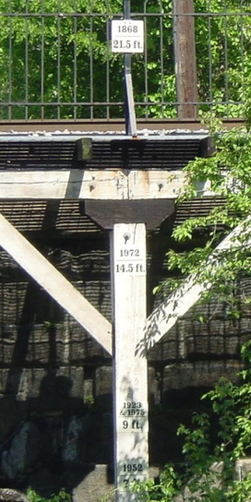 Water level markers on a post, showing measurements from 1868, 1972, 1982, and 1952, surrounded by greenery.