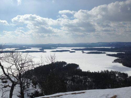 A snowy landscape with a frozen lake, surrounded by islands and trees, under a cloudy sky.