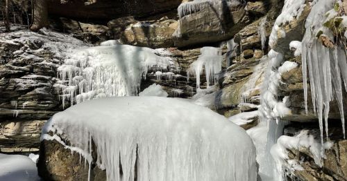 Icicles hang from rocky cliffs and a frozen waterfall, surrounded by snow-covered rocks and trees.