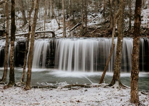 A serene waterfall cascades over rocks, surrounded by trees in a snowy forest setting.