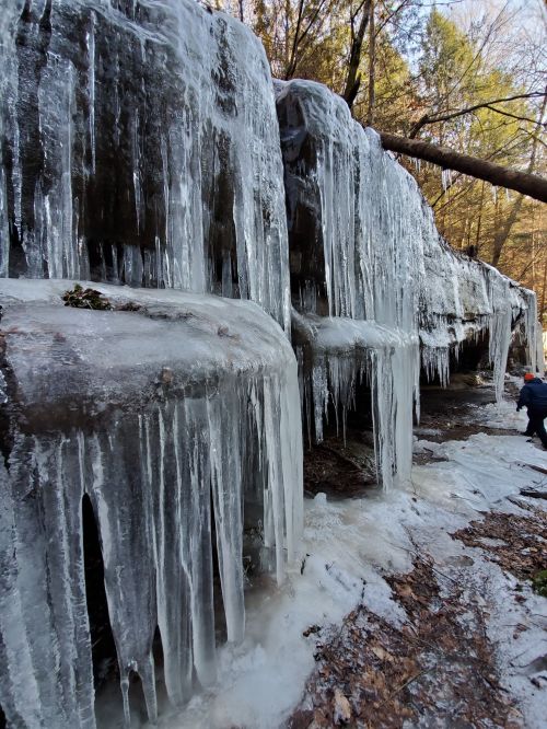 Icicles hang from rocky formations in a forest, with a person in the background near the icy landscape.