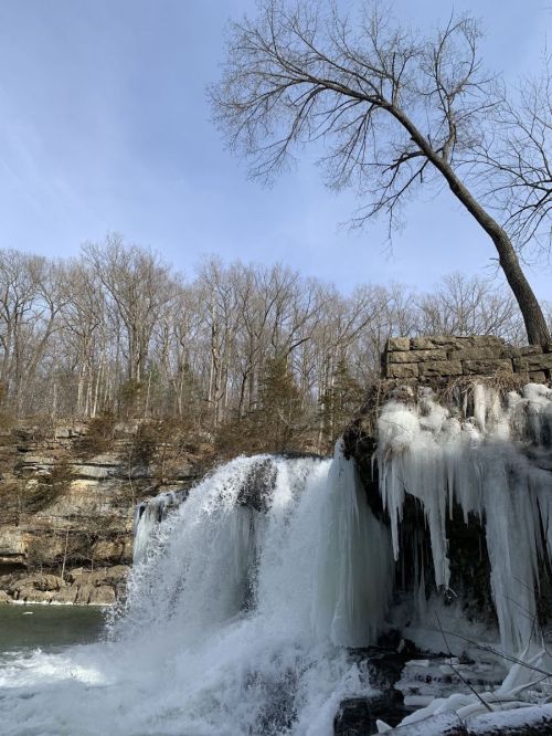 A frozen waterfall surrounded by bare trees and rocky cliffs under a clear blue sky.