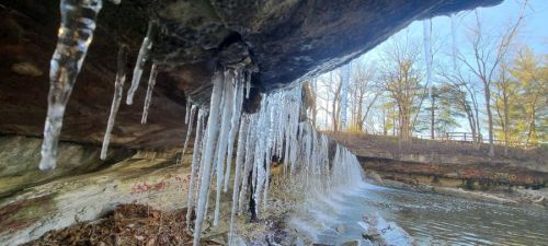 Icicles hang from a rocky overhang above a flowing stream, surrounded by trees in a winter landscape.