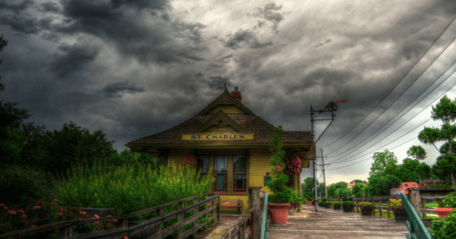 Historic St. Charles train station under a dramatic, cloudy sky, surrounded by greenery and a wooden walkway.