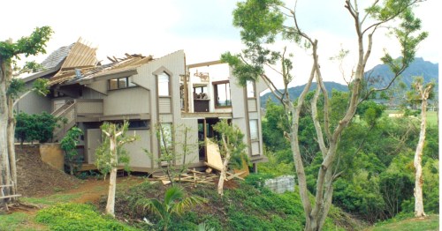 A damaged house with a missing roof, partially collapsed, surrounded by trees and hills in the background.