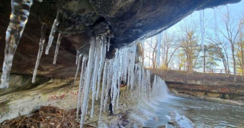Icicles hang from a rocky overhang above a flowing stream, surrounded by trees in a winter landscape.