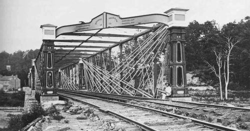 Historic metal bridge with intricate supports, spanning railway tracks, surrounded by trees and a rural landscape.
