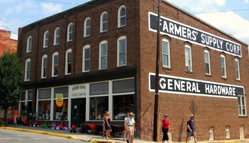 A brick building with "Farmers' Supply Corp General Hardware" painted on the side, featuring people walking by.
