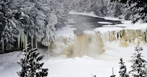 A snowy landscape featuring a waterfall surrounded by frosted trees and icy riverbanks. Steam rises from the water.