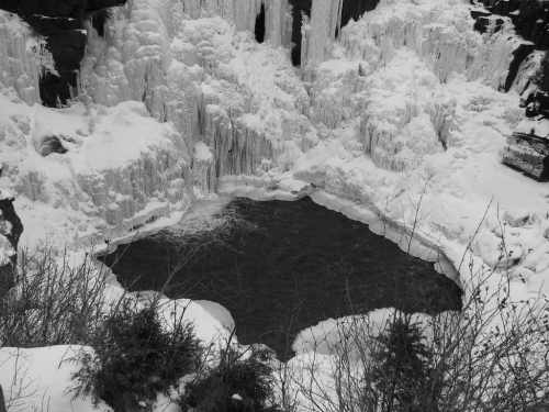 A frozen waterfall surrounded by snow and ice, with a dark pool of water at the center.