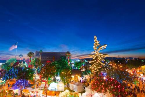 A vibrant holiday market illuminated with colorful lights under a twilight sky, featuring festive decorations and trees.