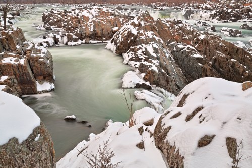 A snowy landscape with rocky riverbanks and flowing water, creating a serene winter scene.
