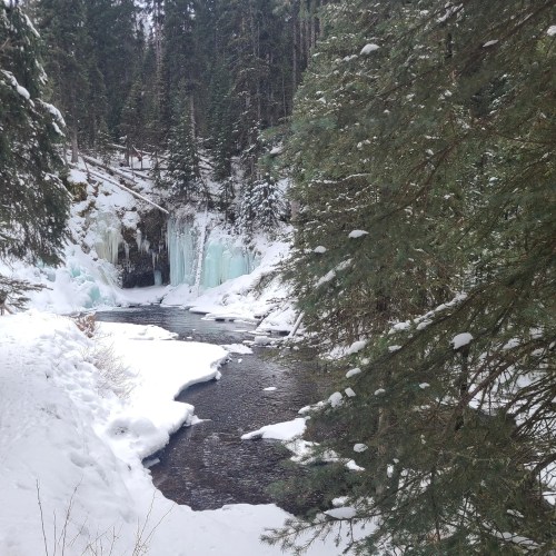 A serene winter scene featuring a frozen waterfall surrounded by snow-covered trees and a flowing river.