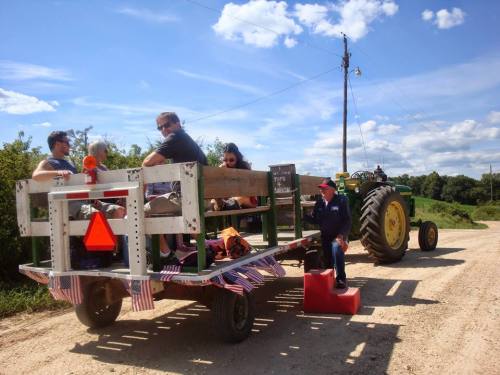 A group of people sitting on a decorated trailer being pulled by a tractor on a dirt road under a blue sky.