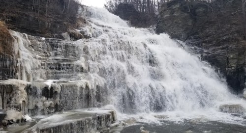 A cascading waterfall flows over rocky cliffs, surrounded by bare trees and icy patches.