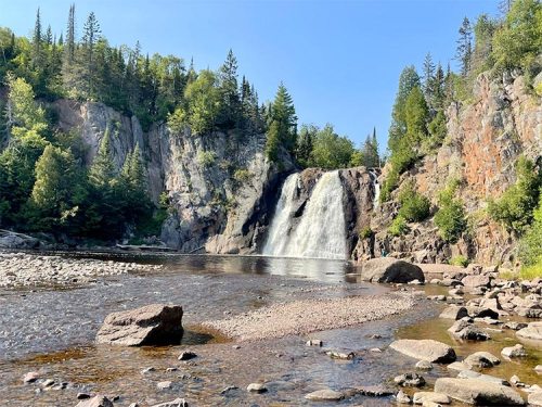 A serene waterfall cascades into a calm river, surrounded by lush greenery and rocky cliffs under a clear blue sky.