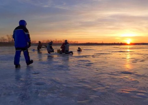 A group of people ice fishing on a frozen lake at sunset, with a colorful sky and silhouettes against the ice.