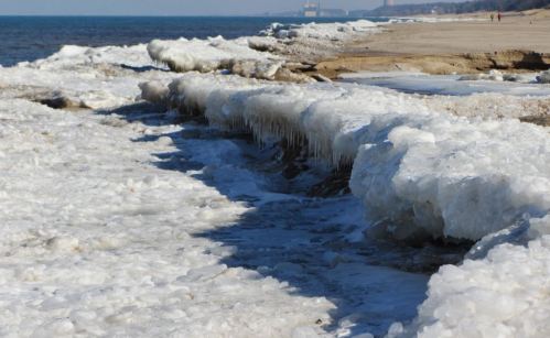 A frozen shoreline with ice formations along the water's edge and a clear blue sky in the background.