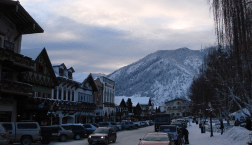 A snowy mountain town with charming buildings, parked cars, and a backdrop of snow-covered mountains under a cloudy sky.