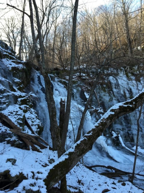 A frozen waterfall surrounded by snow-covered rocks and bare trees in a winter landscape.