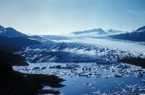 A vast glacier flows into a serene blue lake, surrounded by mountains under a clear sky. Icebergs float in the water.