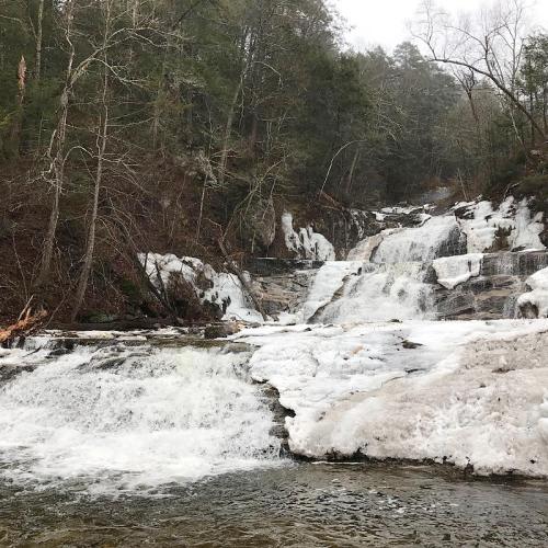 A cascading waterfall surrounded by trees, with patches of ice and snow along the rocky banks.