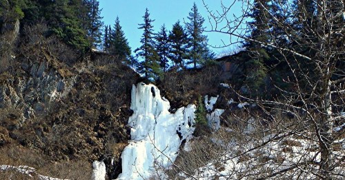 A frozen waterfall cascading down a rocky cliff, surrounded by evergreen trees and a clear blue sky.