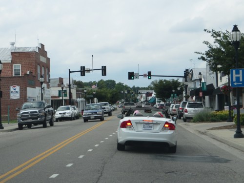 A street view of a small town with traffic lights, parked cars, and storefronts lining the road under a cloudy sky.
