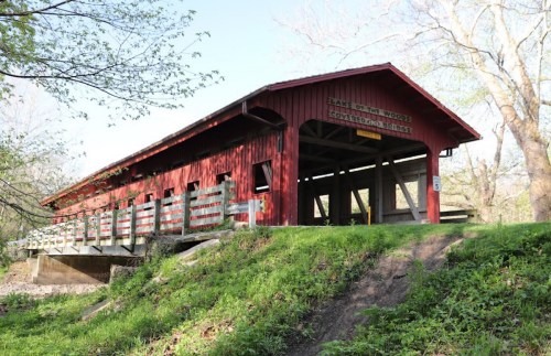 A red covered bridge with wooden railings, surrounded by greenery and trees, spanning a small creek.